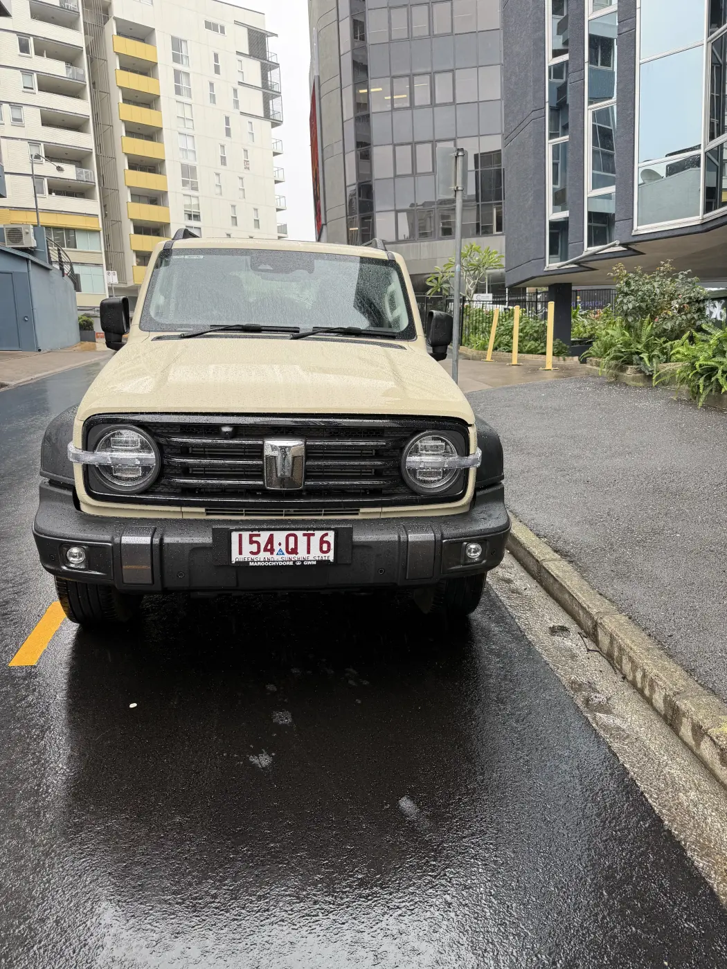 Front view of a cream GWM Tank 300 PHEV parked on a wet Brisbane street, headlights and round grille visible.